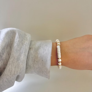 May include: White and gold beaded bracelet. The bracelet features flat, white beads interspersed with small, gold-colored beads. The bracelet is worn on a wrist, with a gray long-sleeved shirt in the background.