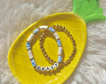 May include: Two bracelets rest on a yellow pineapple-shaped dish. One bracelet is gold, the other is white with blue accents and the word "COAST". The dish is on a white, fluffy surface.