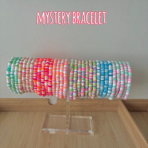 May include: Two stacks of colorful beaded bracelets on a clear display stand. The bracelets are in shades of green, pink, orange, and blue. The text "MYSTERY BRACELET" is displayed above the bracelets.