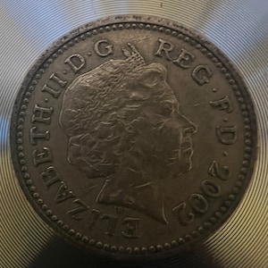 May include: A silver British one pound coin featuring a profile of Queen Elizabeth II with the inscription "ELIZABETH II D.G. REG. F.D. 2002".