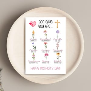 May include: A white card with the text "GOD SAYS YOU ARE..." and "HAPPY MOTHER'S DAY!" is displayed on a wooden tray. The card features a pink heart, a gold cross, and watercolor flowers with words like "Loved" and "Beautiful".