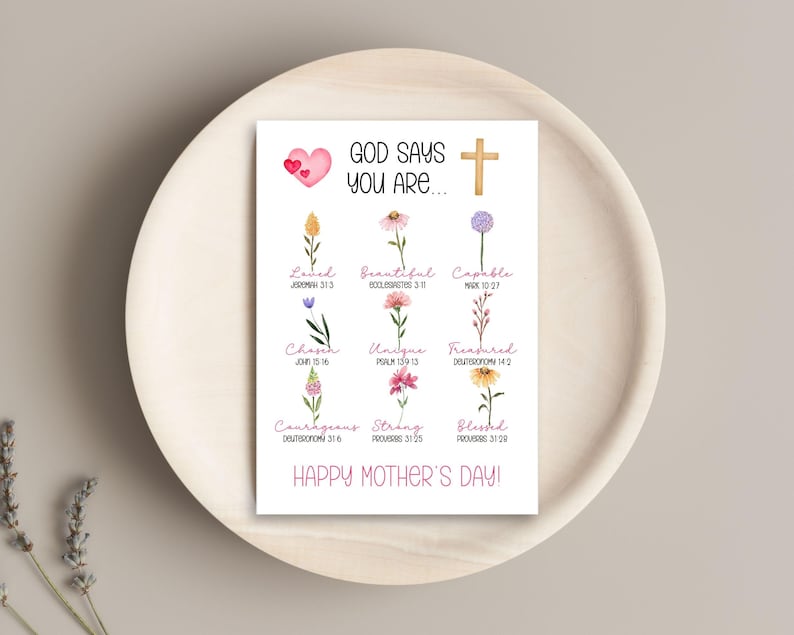 May include: A white card with the text "GOD SAYS YOU ARE..." and "HAPPY MOTHER'S DAY!" is displayed on a wooden tray. The card features a pink heart, a gold cross, and watercolor flowers with words like "Loved" and "Beautiful".