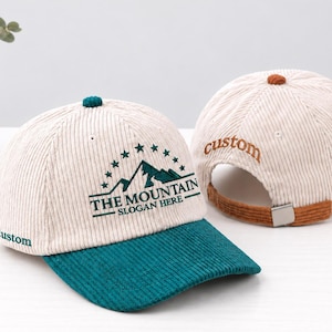 May include: Two corduroy baseball caps in cream with teal and brown accents. The front cap features a teal mountain graphic with stars and the text THE MOUNTAIN SLOGAN HERE. The side and back of the caps feature the embroidered word custom.