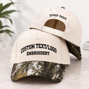 May include: Two cream-coloured baseball caps with camouflage-patterned brims. The front cap features black embroidered text reading CUSTOM TEXT/LOGO EMBROIDERY. The back cap shows a white plastic snapback closure and the black text your text.