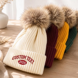 Puede incluir: Una fila de cuatro gorros de punto acanalado, cada uno rematado con un gran pompón de piel sintética. El gorro color crema del frente presenta el texto rojo bordado CUSTOM TEXT y el número 2026. Los otros gorros son burdeos, amarillo y verde oscuro.