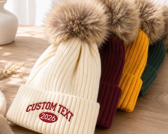 May include: A row of four ribbed knit beanies, each topped with a large faux fur pom-pom. The front cream-colored beanie features the embroidered red text CUSTOM TEXT and the number 2026. The other beanies are solid burgundy, yellow, and dark green.