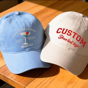 Può includere: Due cappellini da baseball con visiere curve sono mostrati su una superficie di legno. Uno è azzurro con un logo martini e la scritta "CUSTOM LOGO EST. 2026 Martini Club." L'altro è bianco sporco con la scritta rossa "CUSTOM Your text logo."