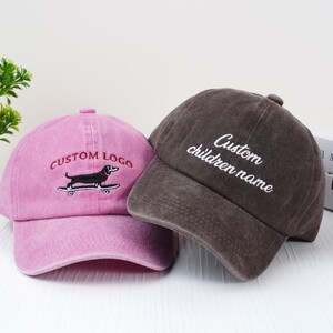 May include: Two baseball caps. One is pink with "CUSTOM LOGO" embroidered above a dachshund on a skateboard. The other is brown with "Custom children name" embroidered on the front. Both caps have a curved brim.
