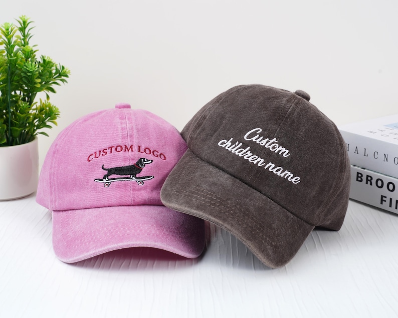 May include: Two baseball caps. One is pink with "CUSTOM LOGO" embroidered above a dachshund on a skateboard. The other is brown with "Custom children name" embroidered on the front. Both caps have a curved brim.