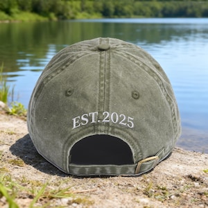 May include: An olive green baseball cap with the text "EST. 2025" embroidered in white. The cap has a curved peak and a brass-coloured adjustable buckle. The cap is resting on a rock near a lake.