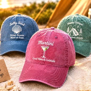 May include: Three baseball caps in blue, magenta, and green. The blue cap features a seashell graphic with the text Salty Soul, Ocean Breeze, Mind at Ease. The magenta cap shows a martini glass with the text Martini, Good Vibes & Cocktails. The green cap displays a tent graphic with the text Happy Camper, Adventure Awaits.