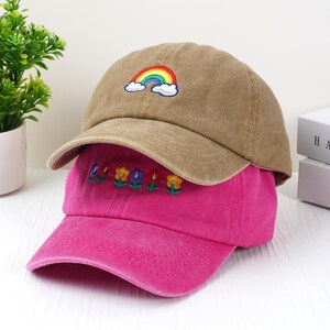 May include: Two baseball caps, one tan and one pink, are stacked. The tan cap has a rainbow and cloud embroidery. The pink cap features embroidered flowers. Both caps have curved brims and are made of a soft fabric.