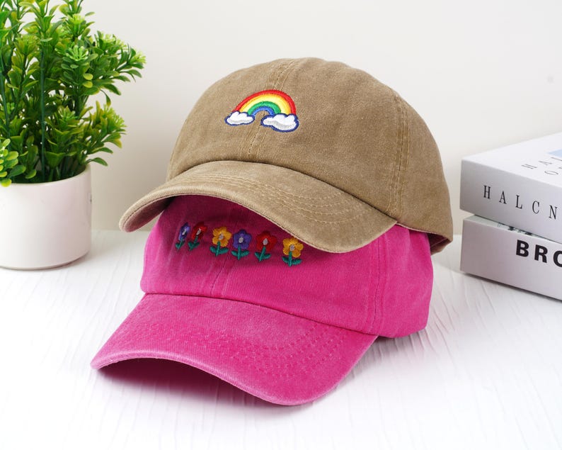 May include: Two baseball caps, one tan and one pink, are stacked. The tan cap has a rainbow and cloud embroidery. The pink cap features embroidered flowers. Both caps have curved brims and are made of a soft fabric.