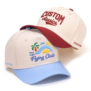 Op de afbeelding: Twee baseballpetten. Eén is crèmekleurig met een rode klep en de woorden "CUSTOM classics" in het rood geborduurd. De andere is crèmekleurig met een lichtblauwe klep en de woorden "Flying Club" in het blauw geborduurd.
