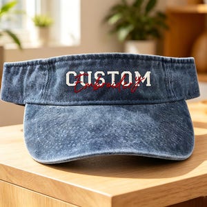 Op de afbeelding: Een blauwe denim vizier met de tekst "CUSTOM Embroidery" in wit en rood. De vizier ligt op een houten oppervlak.