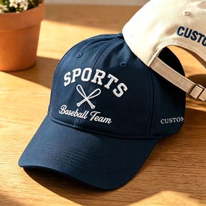 May include: Two baseball caps. One is navy blue with "SPORTS Baseball Team" in white lettering and crossed baseball bats. The other is beige with "CUSTOM" embroidered in blue. Both have adjustable straps.