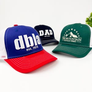 May include: Three trucker hats with different designs. The first hat is blue with white mesh, red brim, and the text "dbla est. 2024". The second hat is navy blue with white mesh and the text "DAD est. 2024". The third hat is green with white mesh and the text "THE MOUNTAIN - SLOGAN HERE".
