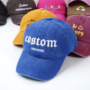 May include: A collection of baseball caps in various colors, including blue, yellow, and maroon. The blue cap in the foreground has the word "custom" embroidered in white. Other caps feature text like "Custom children name" and "Be happy".