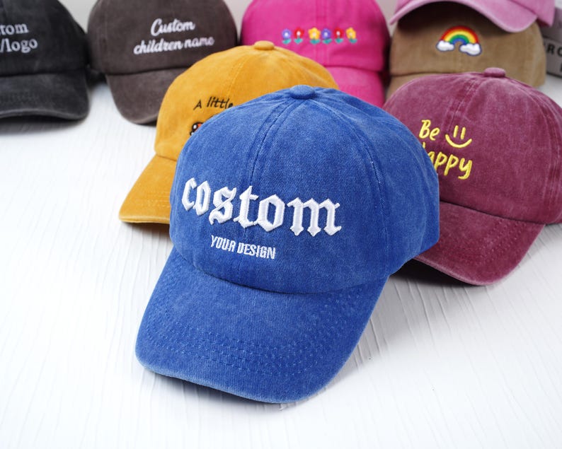 May include: A collection of baseball caps in various colors, including blue, yellow, and maroon. The blue cap in the foreground has the word "custom" embroidered in white. Other caps feature text like "Custom children name" and "Be happy".