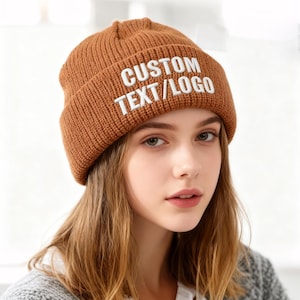 May include: A brown knitted beanie hat with the text "CUSTOM TEXT/LOGO" in white. The hat is made of a ribbed knit fabric. The person wearing the hat has long, light brown hair.