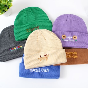 May include: A selection of knitted beanies in various colours. The beanies are in green, grey, cream, purple, blue, and brown. Each beanie has embroidered designs or text, including flowers, a dog, the word "smile", and "Best dad".
