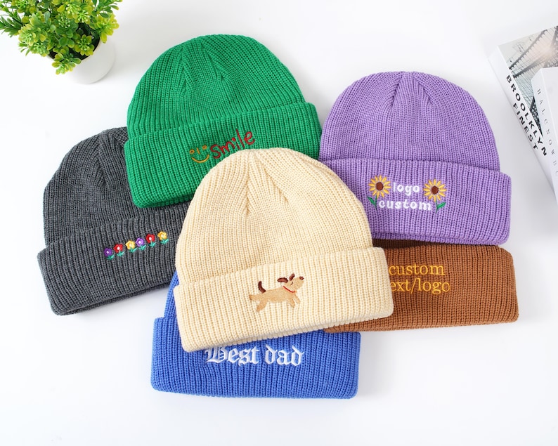 May include: A collection of colorful knit beanies. The beanies are in various colors, including green, gray, cream, purple, blue, and brown. Each beanie features embroidered designs or text, such as flowers, a dog, the word "smile", and "Best dad".