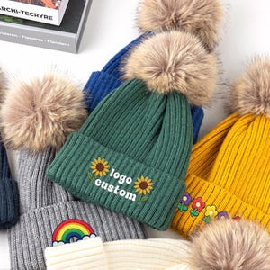 Peut inclure: Une collection de bonnets en tricot côtelé de différentes couleurs, notamment bleu foncé, gris, vert et jaune, chacun surmonté d'un pompon en fausse fourrure. Le bonnet vert présente des tournesols brodés et le texte logo custom en blanc.
