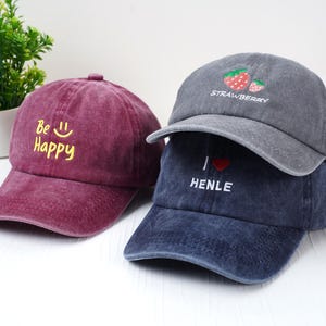 May include: Three baseball caps in shades of burgundy, gray, and navy blue. The burgundy cap has the text "Be Happy" in yellow. The gray cap features a strawberry design and the word "Strawberry". The navy cap says "I ❤️ HENLE".