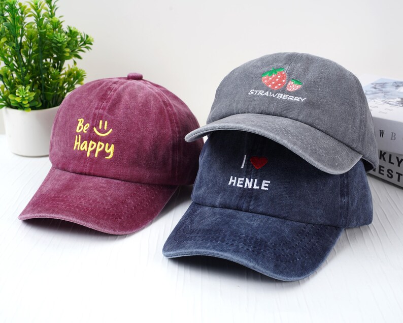 May include: Three baseball caps in shades of burgundy, gray, and navy blue. The burgundy cap has the text "Be Happy" in yellow. The gray cap features a strawberry design and the word "Strawberry". The navy cap says "I ❤️ HENLE".