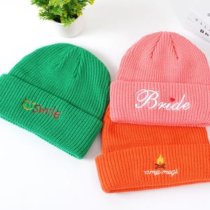 May include: Three knitted beanies in green, coral, and orange. The green beanie has "smile" embroidered in yellow and red. The coral beanie says "Bride" in white script. The orange beanie has "camp megs" and a campfire embroidered in yellow.