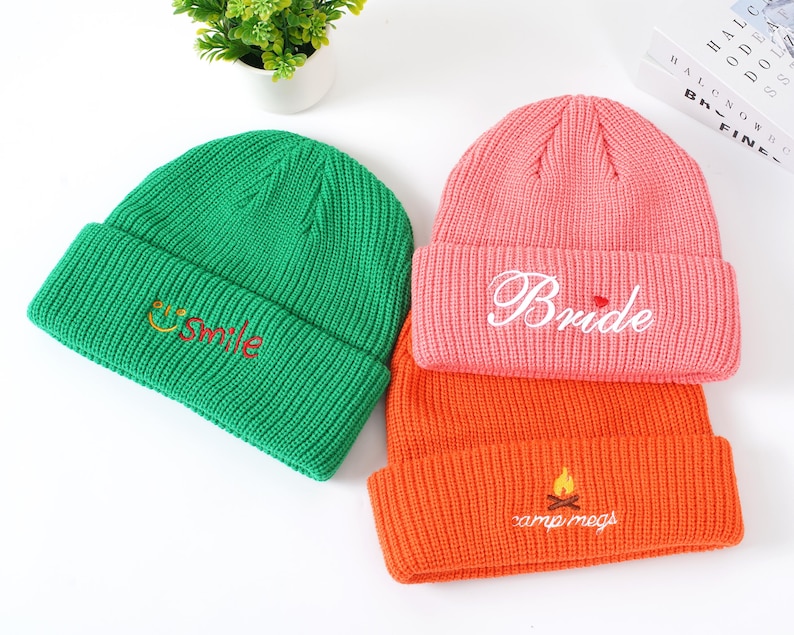 May include: Three knitted beanies in green, coral, and orange. The green beanie has "smile" embroidered in yellow and red. The coral beanie says "Bride" in white script. The orange beanie has "camp megs" and a campfire embroidered in yellow.