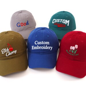 May include: Five baseball caps in different colours: grey, green, blue, red, and olive green. Each cap has embroidered text or designs, including "Good", "Custom Classics", "Custom Embroidery", a cherry, and flowers with "Wonderful day".