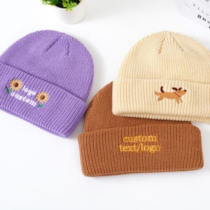 May include: Three knit beanies in purple, tan, and brown. The purple beanie has embroidered sunflowers and the text "logo custom". The tan beanie features a dog graphic. The brown beanie has the text "custom text/logo".