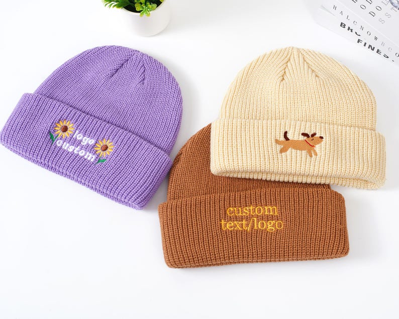 May include: Three knit beanies in purple, tan, and brown. The purple beanie has embroidered sunflowers and the text "logo custom". The tan beanie features a dog graphic. The brown beanie has the text "custom text/logo".