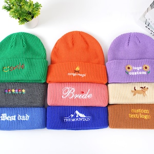 May include: A collection of colorful knit beanies. The beanies are in green, orange, purple, gray, pink, blue, and tan. Some beanies have embroidered text or designs, including "Smile", "Bride", "Best dad", and "custom text/logo".