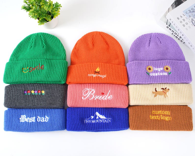 May include: A collection of colorful knit beanies. The beanies are in green, orange, purple, gray, pink, blue, and tan. Some beanies have embroidered text or designs, including "Smile", "Bride", "Best dad", and "custom text/logo".