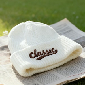 Peut inclure: Un bonnet en tricot blanc avec le mot "classic" brodé en script marron. Le chapeau est plié au bord et repose sur un journal. L'arrière-plan est une pelouse verte floue.