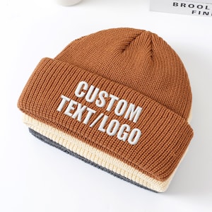 Könnte beinhalten: Eine braune Strickmütze mit umgeschlagenem Rand. Der Rand zeigt den gestickten Text "CUSTOM TEXT/LOGO" in Weiß. Die Mütze hat ein mehrschichtiges Design mit einem cremefarbenen und grauen Band am Saum.