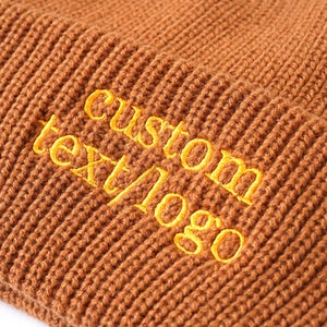 May include: Close-up of a brown knit beanie with the embroidered text "custom text/logo" in yellow. The beanie has a ribbed texture and is made of a soft, warm material. This hat is a customizable accessory.
