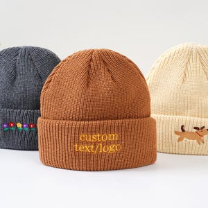 May include: Three knit beanies in a studio setting. A dark gray beanie with embroidered flowers, a brown beanie with "custom text/logo" in yellow, and a cream beanie with a dog embroidery. All have folded brims.