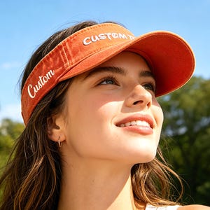 Könnte beinhalten: Ein orangefarbenes Visier mit dem Wort "Custom" in Weiß gestickt. Das Visier wird von einer Person im Freien an einem sonnigen Tag getragen. Der Hintergrund umfasst Bäume und einen blauen Himmel.