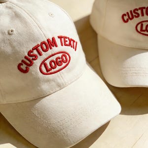 Könnte beinhalten: Zwei cremefarbene Baseballkappen mit gebogenen Schirmen. Jede Kappe hat den roten Schriftzug "CUSTOM TEXT/LOGO". Die Kappen sind aus weichem Stoff und wahrscheinlich verstellbar.