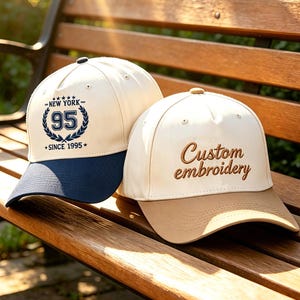 May include: Two baseball caps with curved peaks sit on a wooden bench. One is cream-coloured with a navy blue peak and the text "NEW YORK 95 SINCE 1995." The other is cream-coloured with a tan peak and the text "Custom embroidery."