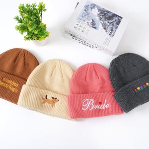 May include: Four knitted beanies in various colors: brown with custom text, cream with a dog, pink with "Bride" script, and gray with floral embroidery. The beanies are displayed on a white surface.