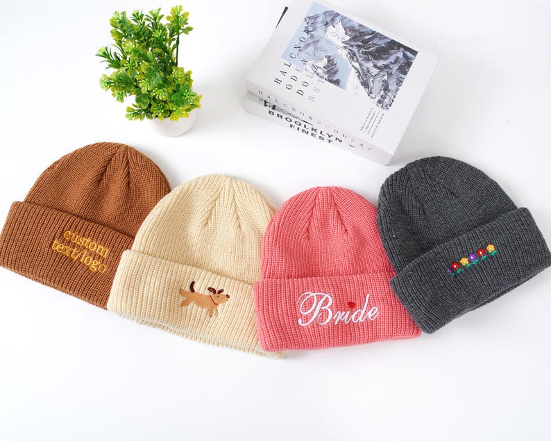 May include: Four knitted beanies in various colors: brown with custom text, cream with a dog, pink with "Bride" script, and gray with floral embroidery. The beanies are displayed on a white surface.