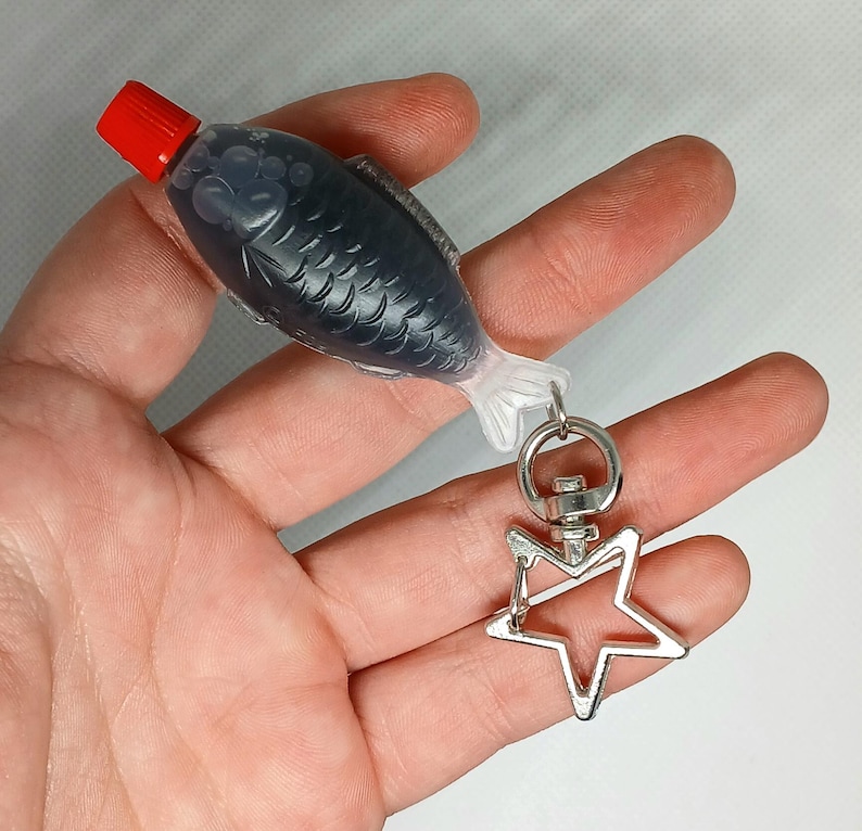 Pu&ograve; includere: Un portachiavi originale con una bottiglia di salsa di soia a forma di pesce con tappo rosso. La bottiglia grigio scuro ha un motivo a squame di pesce ed &egrave; attaccata a un ciondolo a forma di stella argentata.