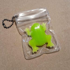 以下が含まれることがあります： ボールチェーンキーホルダー付きの透明なプラスチックポーチに入った、明るい緑色のカエル型のおもちゃ。カエルは光沢のある仕上がりで、鮮やかな緑色です。ポーチは密封されており、透明なのでカエルが見えます。