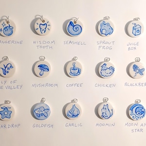 May include: A set of 15 ceramic charms with blue and white painted designs. The charms feature various images, including a tangerine, a wisdom tooth, a seashell, a sprout frog, a juice box, a lily of the valley, a mushroom, a cup of coffee, a chicken, a blackberry, a star drop, a goldfish, a garlic clove, a Moomin character, and a moon and star.