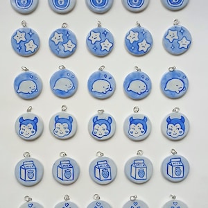 May include: A collection of blue and white ceramic pendants with various designs. The pendants feature illustrations of a jar of jam, stars, a seal, a devil, a milk carton with a strawberry, and a bat. Each pendant has a silver loop for hanging.