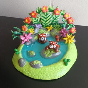 Puede incluir: Una escena en miniatura de arcilla con dos pandas rojos en un estanque azul rodeado de vegetación verde y flores de colores.
